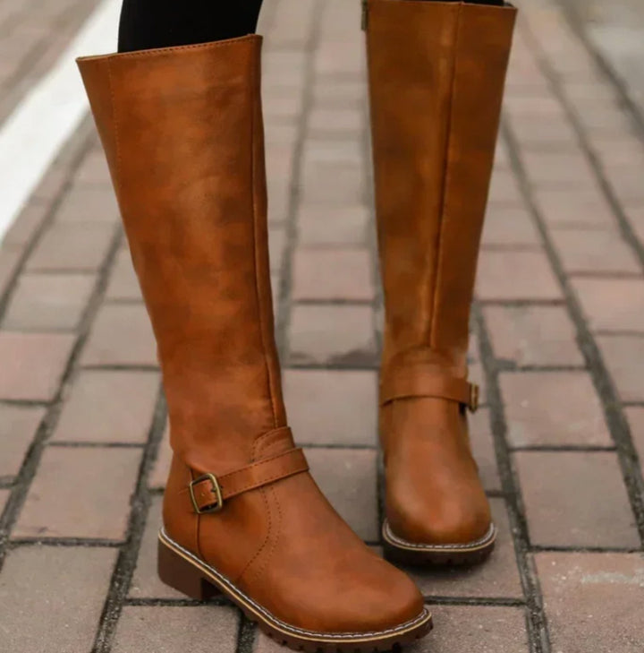 Tan knee high riding boots with an ankle buckle strap and lug sole, worn on a brick path