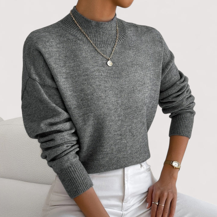 Woman in a grey loose fit knit sweater with a high ribbed neckline and long cuffed sleeves