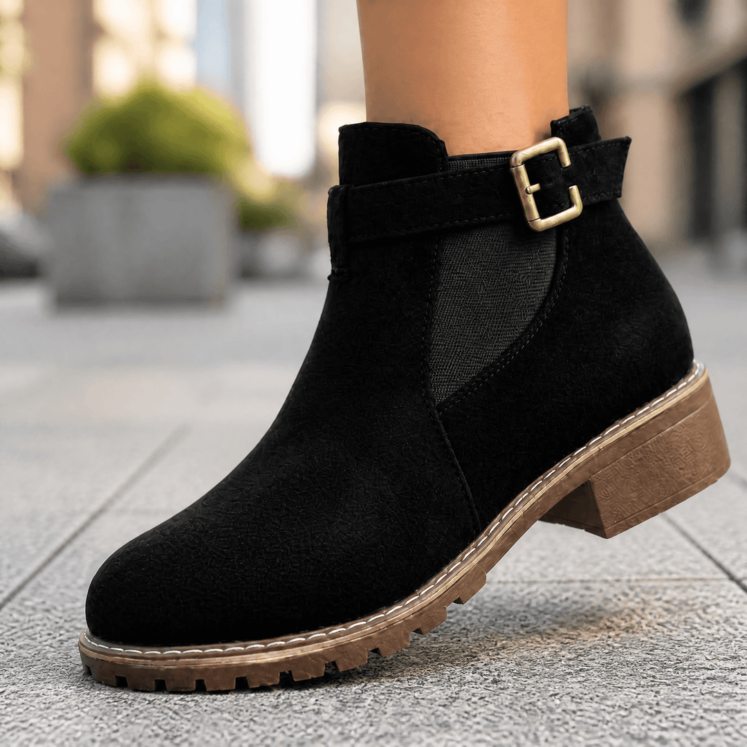 Black suede ankle boot with a gold buckle strap and stacked block heel, worn on a city sidewalk