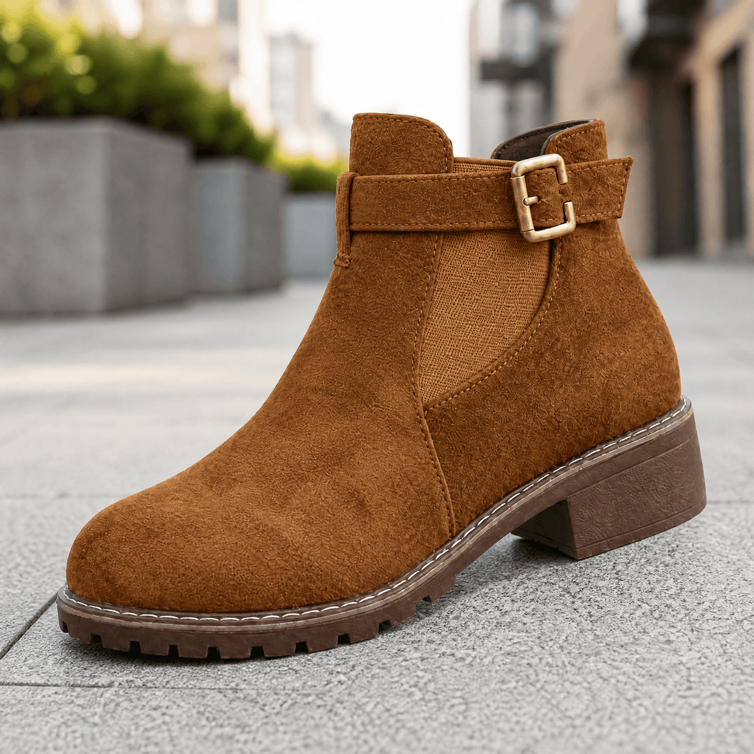 Brown suede ankle boot with a gold buckle strap and stacked heel on pavement