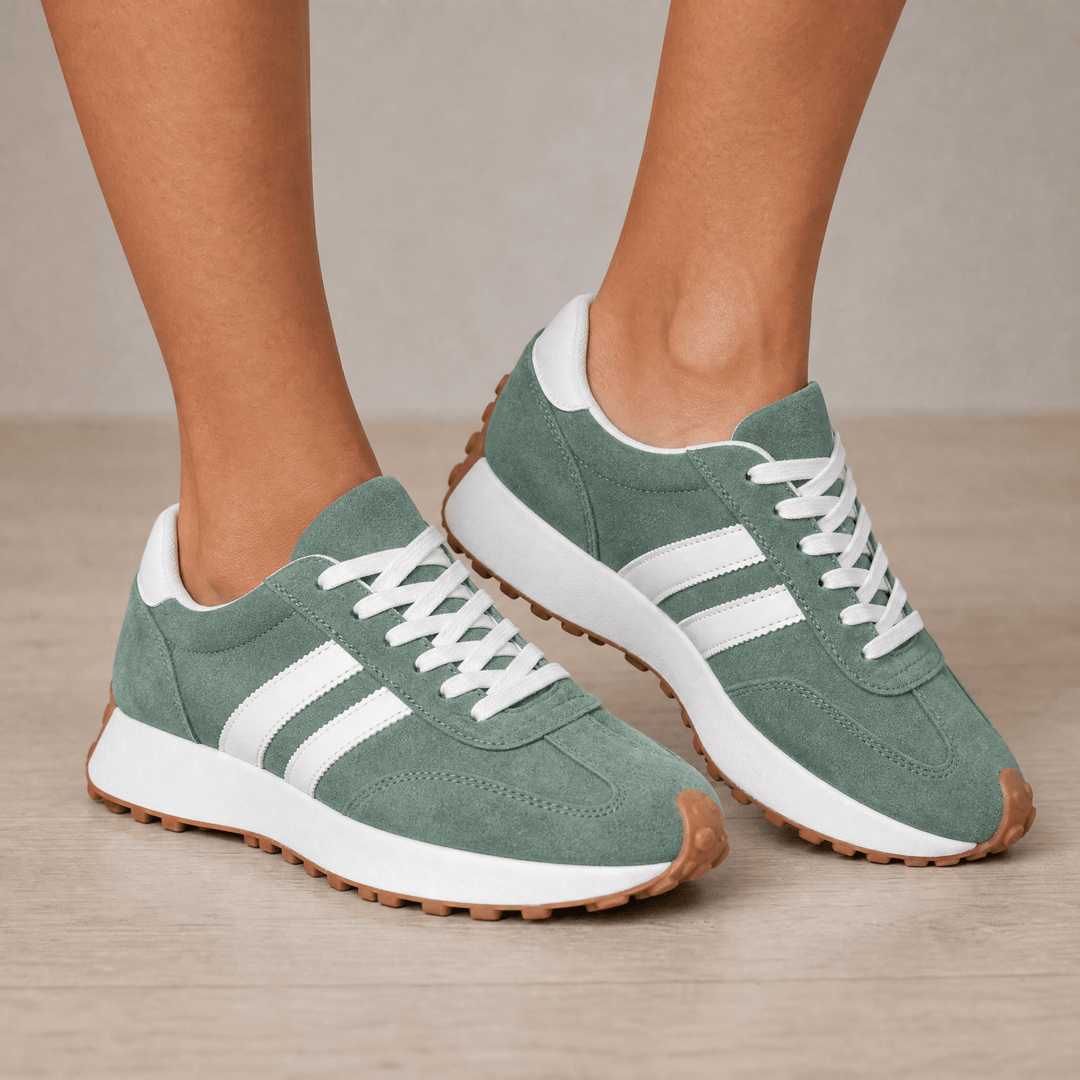 Sage green suede sneakers with white side stripes and a gum sole, worn on a wooden floor