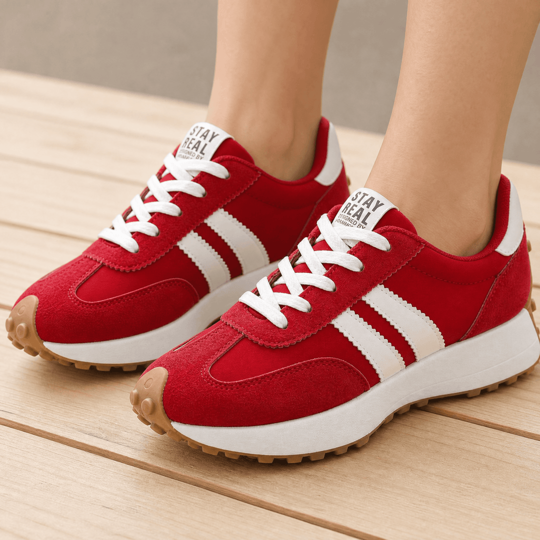 Red suede sneakers with white side stripes, white laces and gum soles, worn on a wooden deck
