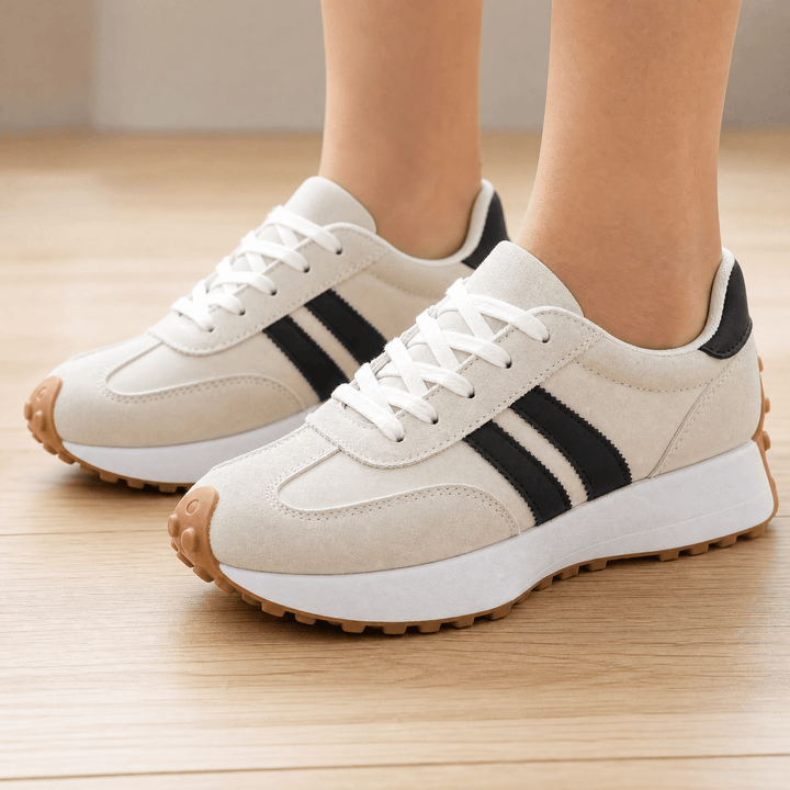 Off white suede sneakers with black side stripes and a gum sole, worn on a wooden floor