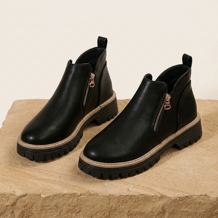 Pair of black ankle boots with a gold side zip and lug sole, standing on a stone ledge