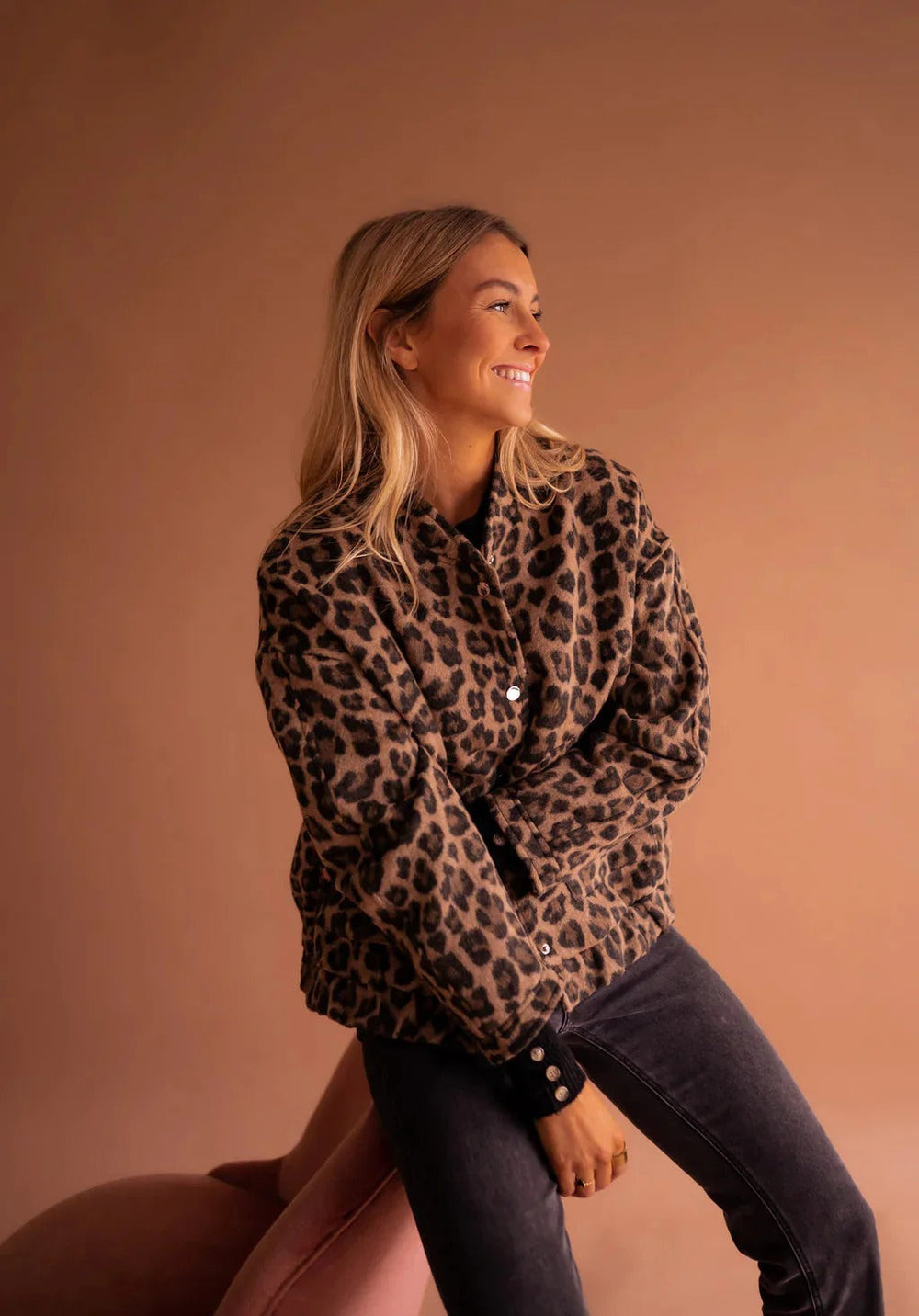 Woman seated in a leopard print bomber jacket and grey jeans