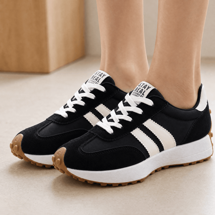 Black suede sneakers with cream side stripes, white laces and gum soles, worn indoors