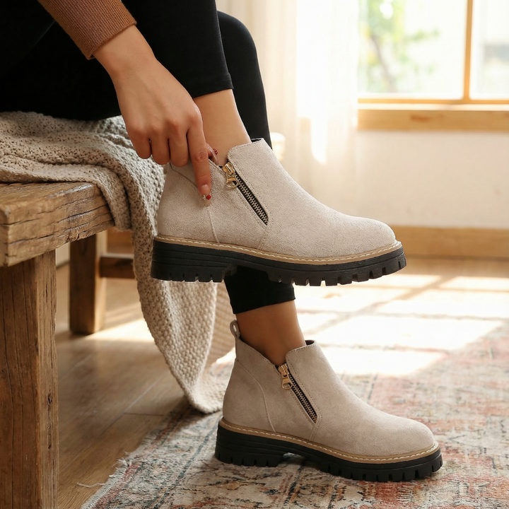 Hand zipping a cream suede chelsea boot beside a knit throw